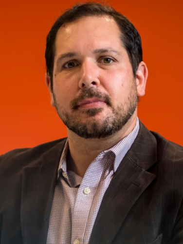 John Wihbey, a light skinned man with short dark hair and short cropped facial hair, looks into the camera wearing a button down shirt and dark colored jacket, in front of a dark orange background.