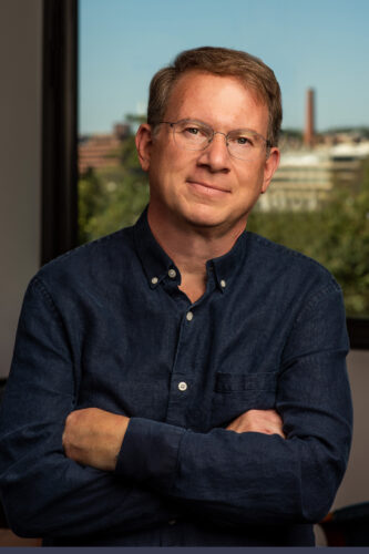 Jeffrey Goldberg wearing glasses and a dark blue button-down shirt stands with his arms crossed in front of a window with a blurred cityscape in the background.