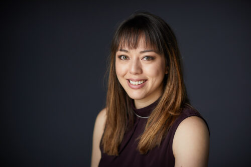 Smiling woman with straight brown hair and bangs, wearing a sleeveless dark top against a dark background.
