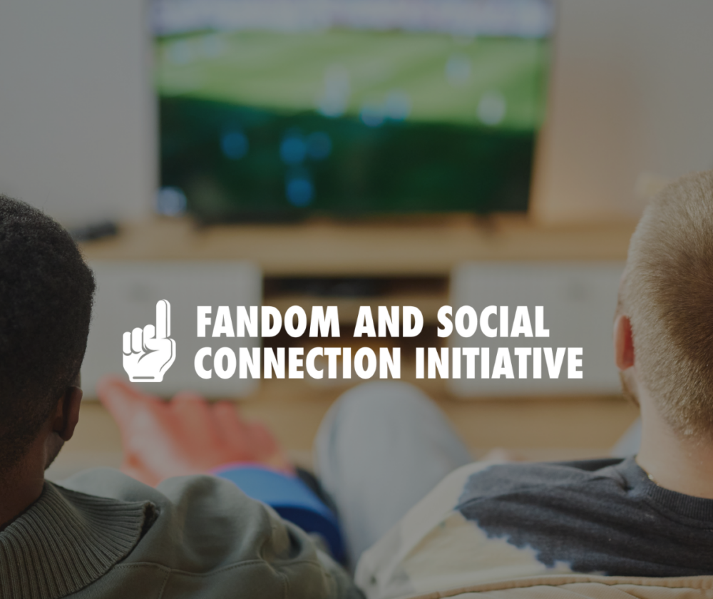 two people sit on a couch watching sports together. the logo for the Fandom and Social Connection Initiative is overlayed in the top left corner.
