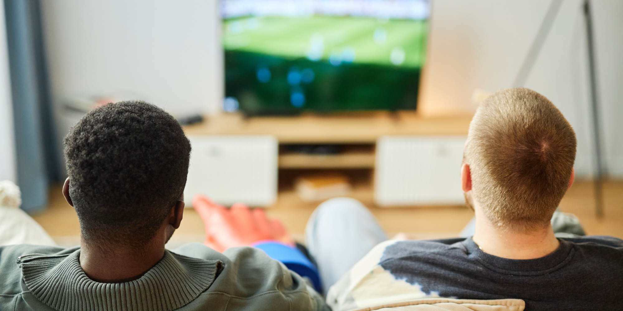 two people with short hair, likely men, viewed from behind while sitting on a couch watching sports on television. One is dark skinned with black hair, and one is light skinned with blonde hair. They sit shoulder to shoulder on the couch, seemingly watching companionably.