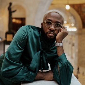 Caleb Okereke, a brown skinned person with short blonde hair and facial hair, wearing a green satin shirt and glasses, leans his chin on his hand