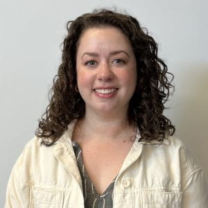 Lisa Thalhamer, a light skinned person with shoulder length brown curly hair, stands looking into the camera wearing a cream colored jacket and green shirt
