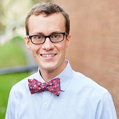 Headshot of a smiling man with a bow tie and glasses in front of a brick wall outside