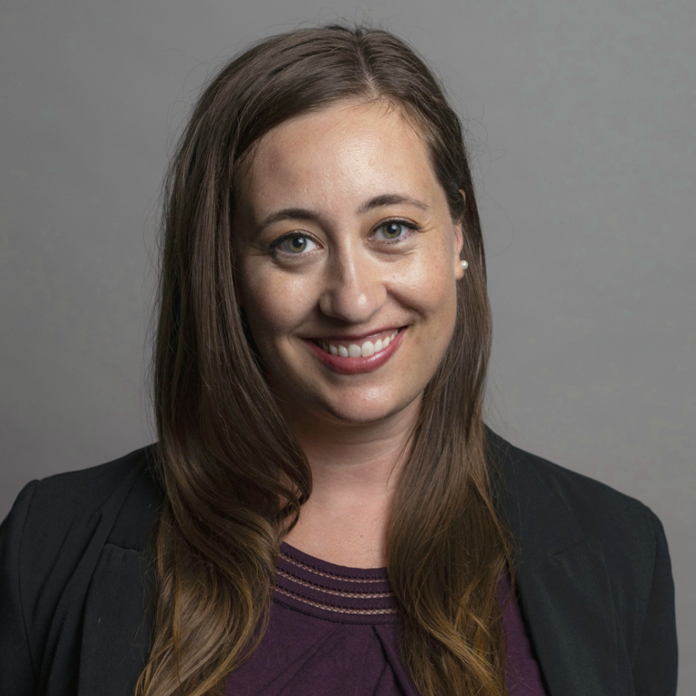 Headshot of a smiling woman with long brown hair wearing a dark blazer and purple top against a gray background.