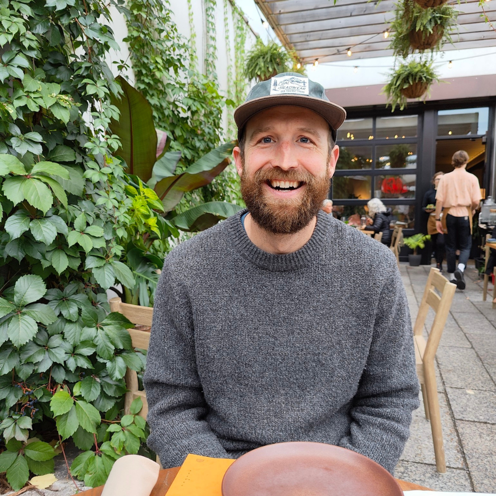 Smiling bearded man wearing a gray sweater and baseball cap, seated at an outdoor restaurant surrounded by greenery.