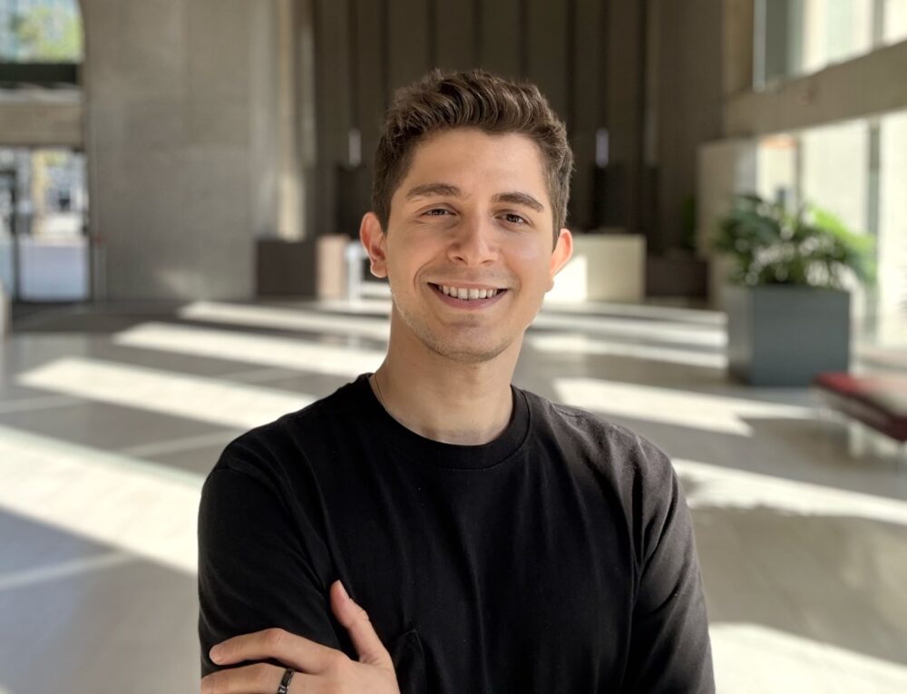 Ata Uslu, a man with short dark hair and light skin smiles while wearing a dark colored shirt, standing inside a large indoor space with light falling across the floor from large windows.