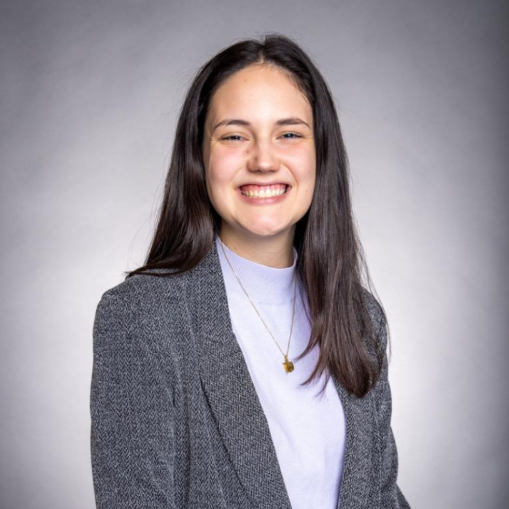 Headshot of a smiling woman with long dark hair, wearing a gray blazer over a light top against a gray background.