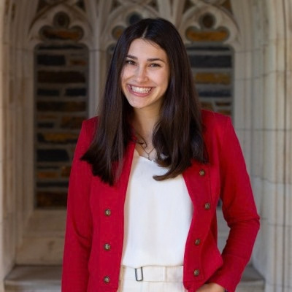 Headshot of a smiling woman with long dark hair, wearing a red blazer over a white top in front of an arched stone building.
