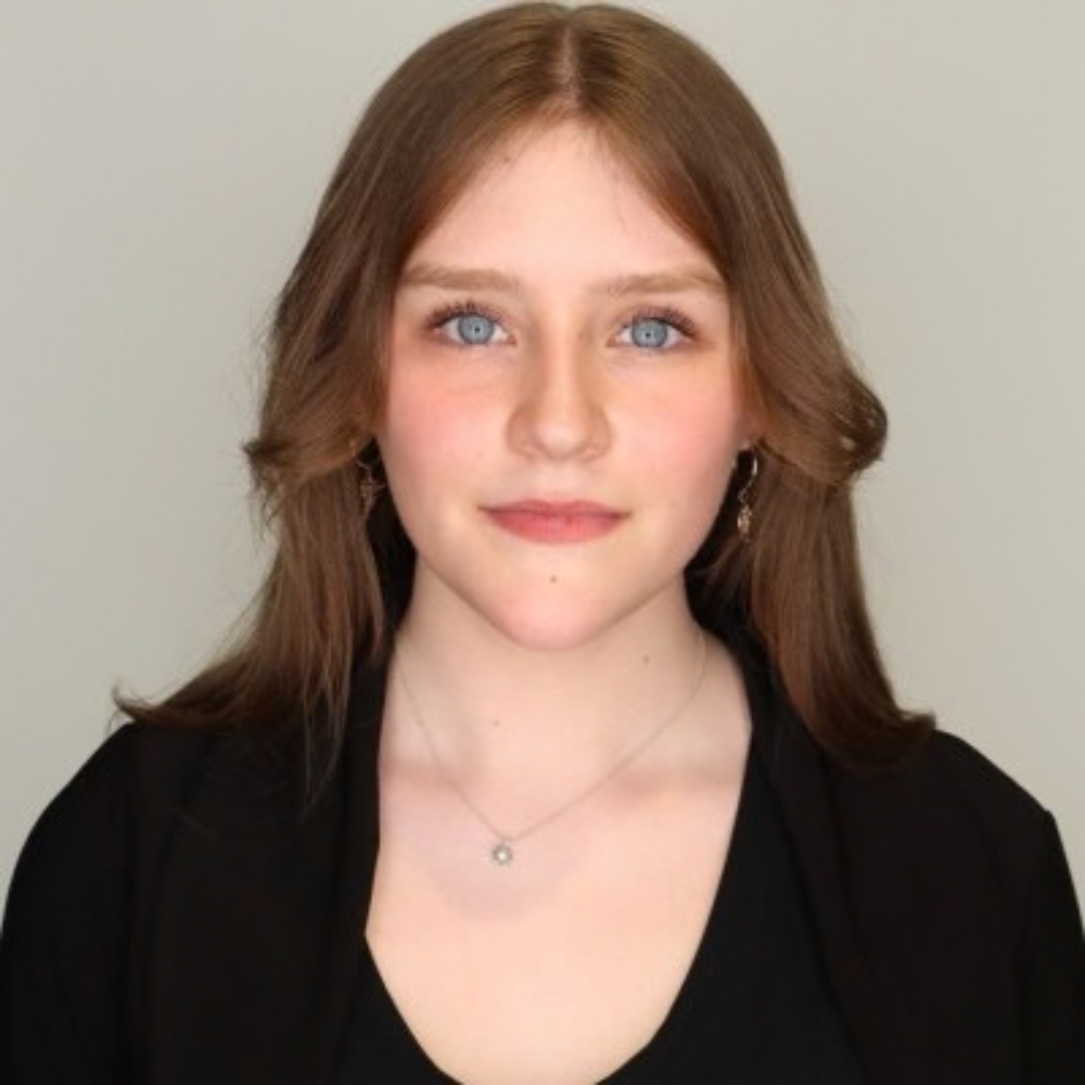 Headshot of a woman with shoulder-length brown hair wearing a black top and a delicate necklace against a light gray background.