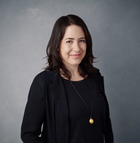 Headshot of Angèle Christin smiling, with shoulder-length dark brown hair, wearing a black blazer and a gold pendant necklace against a gray background.