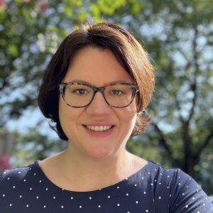 Rebecca Piazza, a light skinned woman with short reddish brown hear wearing glasses and a blue shirt sitting outside near trees