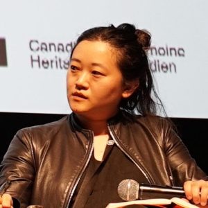 Abby Sun, a woman of Asian descent with dark hair pulled back, sits on a stage with a microphone, wearing a dark leather jacket