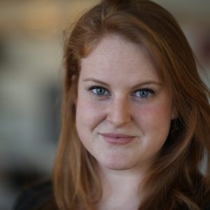 Allison Abbott, a light skinned woman with red hair and blue eyes, looks into the camera with a blurred background behind her
