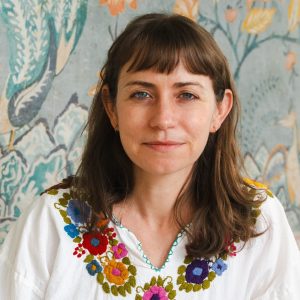 Erin Zimmer Strenio, a light skinned woman with shoulder length brown hair wears a white shirt with embroidered flowers, in front of a decorative mural.