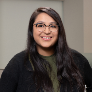 photo of Lauren Faz, a mexican-american woman with long dark brown hair, wearing glasses and a black sweater and green shirt.