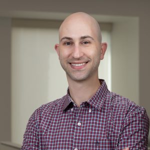 Seth Goldman, a light skinned bald man wearing a short sleeved red checked shirt