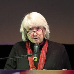 Mary Lampson, a light skinned woman with chin length gray hair, standing at a lecturn