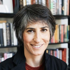 a light skinned woman with short dark hair wearing a black jacket and red shirt, sitting in front of a bookshelf, smiles into the camera