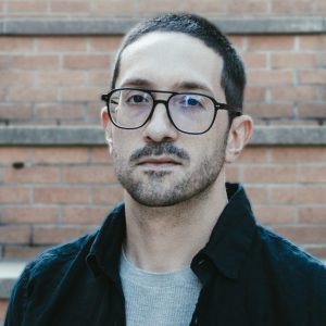 photo of Brian Friedberg, a light skinned man with short dark hair and close-cropped facial hair, wearing black-rimmed glasses.