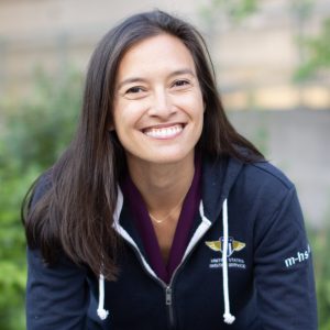 Mina Hsiang, a woman of Asian descent with light skin and long dark hair, smiles towards the camera wearing a blue zip-up sweatshirt with the USDS logo on the front.