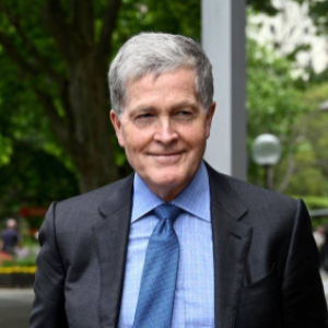 Steven Johnson, a light skinned man with short gray hair wearing a blue suit stands outside near some trees and a building