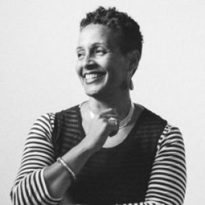 Black and white photo of Tabitha Jackson, a medium skinned woman with short curly dark hair. She is wearing a striped long-sleeved top with a dark overlay on the body.